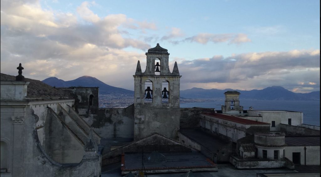 A stone bell tower with bells visible, overlooking the Naples skyline and Mount Vesuvius at dusk