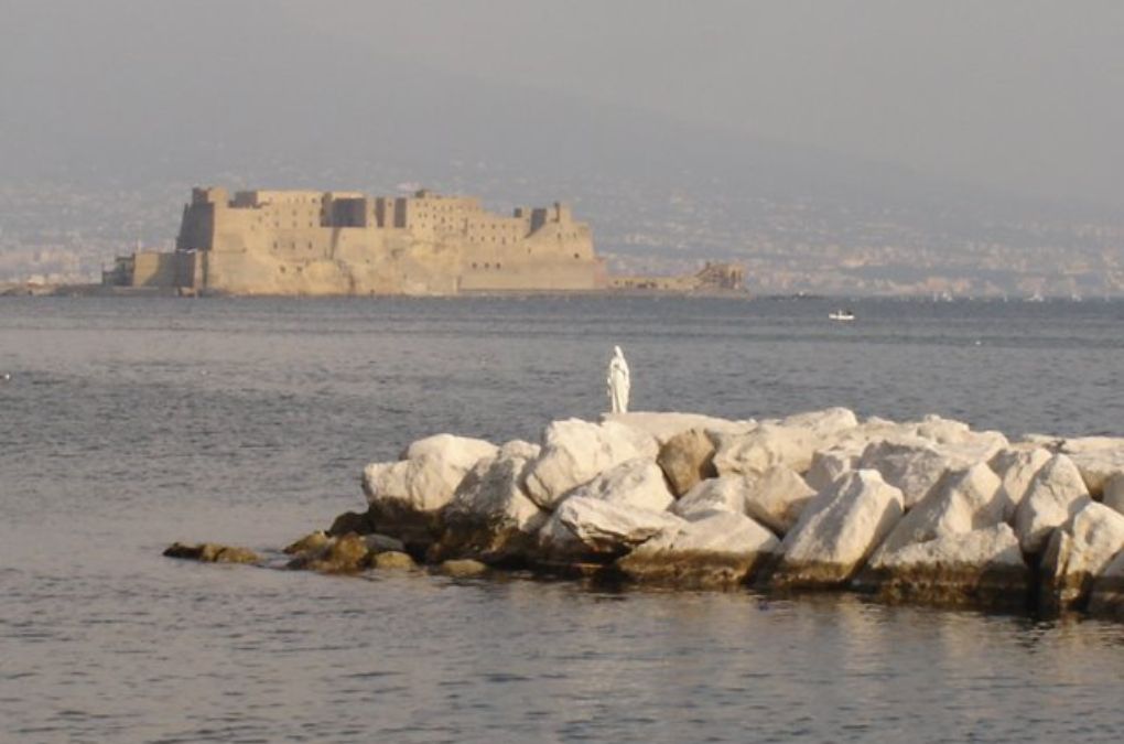 Castel dell'Ovo across the Bay of Naples, with a small statue of the Madonna standing on white rocks in the foreground