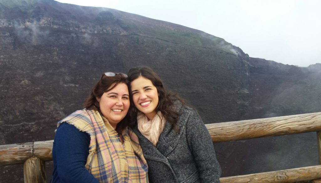 Daniela Pescuma and her sister smiling at the edge of the Vesuvius crater, with the crater wall and drifting steam behind them