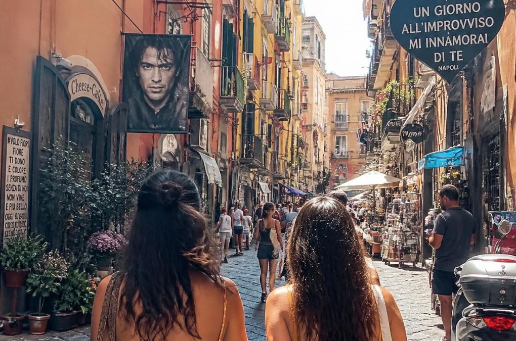 Two friends walking down a busy Naples street beneath a sign reading "Un giorno all'improvviso mi innamorai di te"