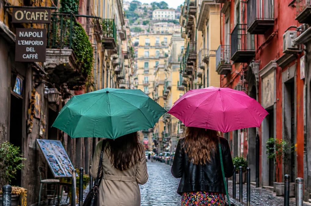 Two friends walking with green and pink umbrellas down a rainy street in Naples