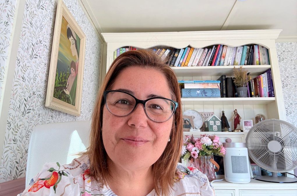 Daniela Pescuma smiling at her desk in her garden writing shed, with bookshelves behind her