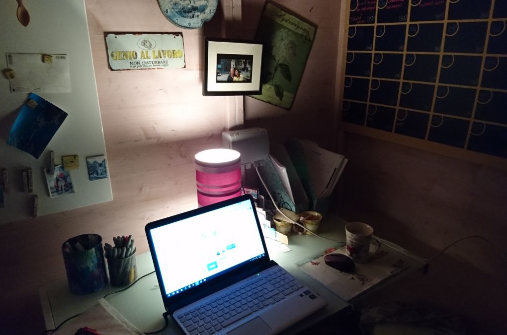 A dimly lit writing desk at night with a laptop, pink lamp, and an Italian "genius at work" sign on the wall