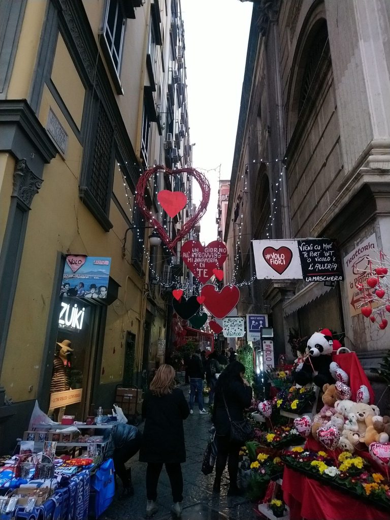 A narrow Naples alley decorated with red hearts and lights for Valentine's Day, with flower stalls below