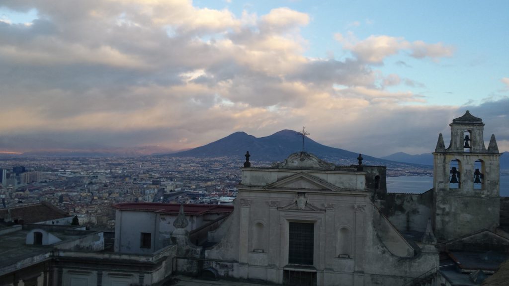 Mount Vesuvius at sunset behind a baroque church facade and bell tower, with the rooftops of Naples and the bay below