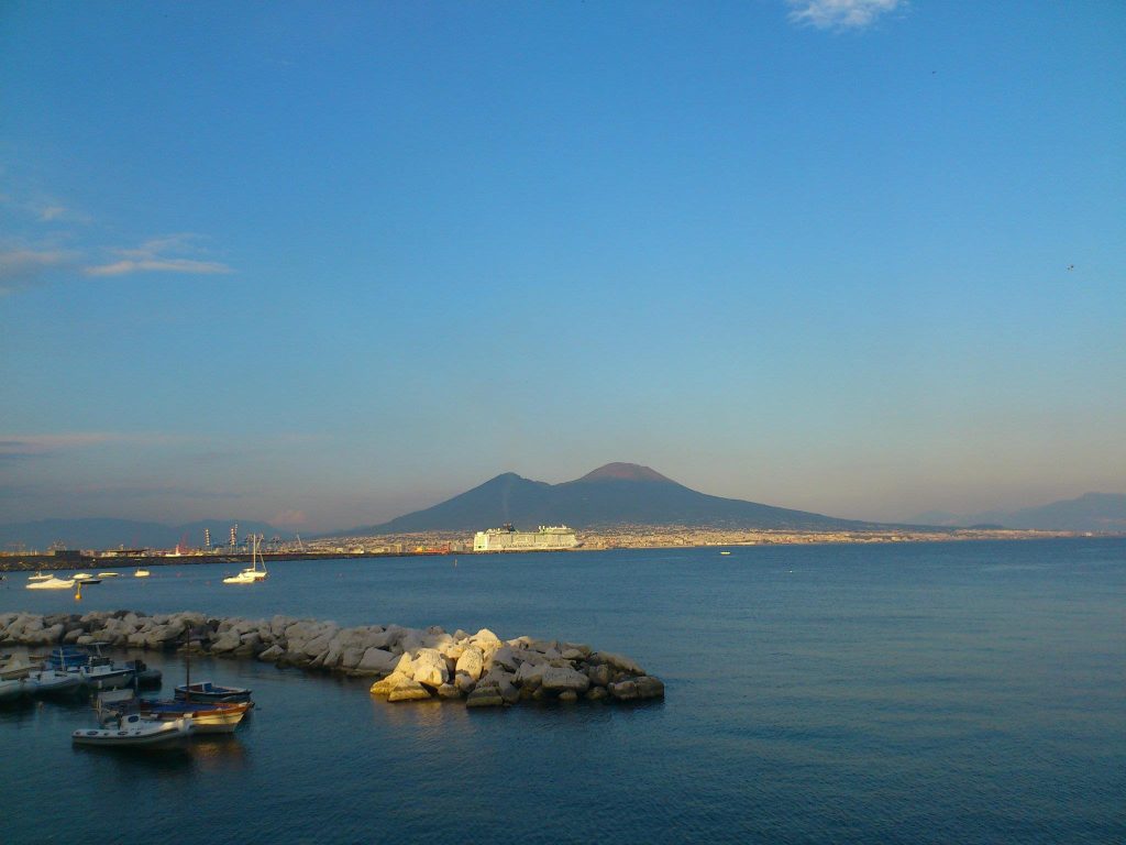Mount Vesuvius across the Bay of Naples at golden hour, with fishing boats moored behind a rock breakwater