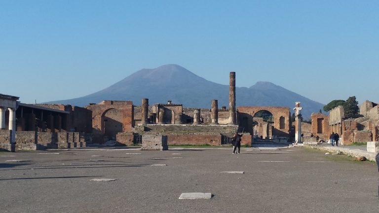 Mount Vesuvius seen across the ruined forum of Pompeii, with columns and brick arches in the foreground under a clear blue sky